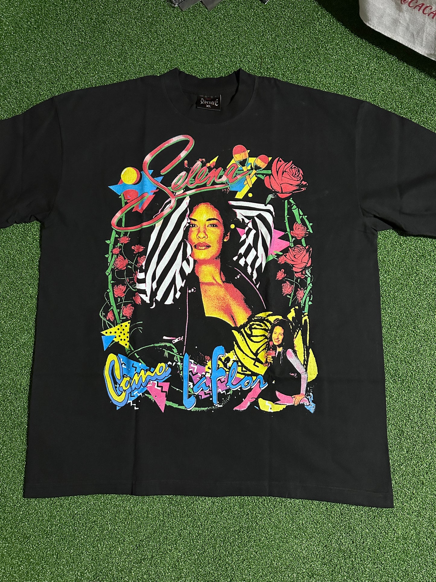 A black long-sleeve graphic t-shirt laid flat on green artificial turf. The shirt features a large, colorful front print with a stylized portrait, roses, and abstract shapes in yellow, pink, blue, and white.