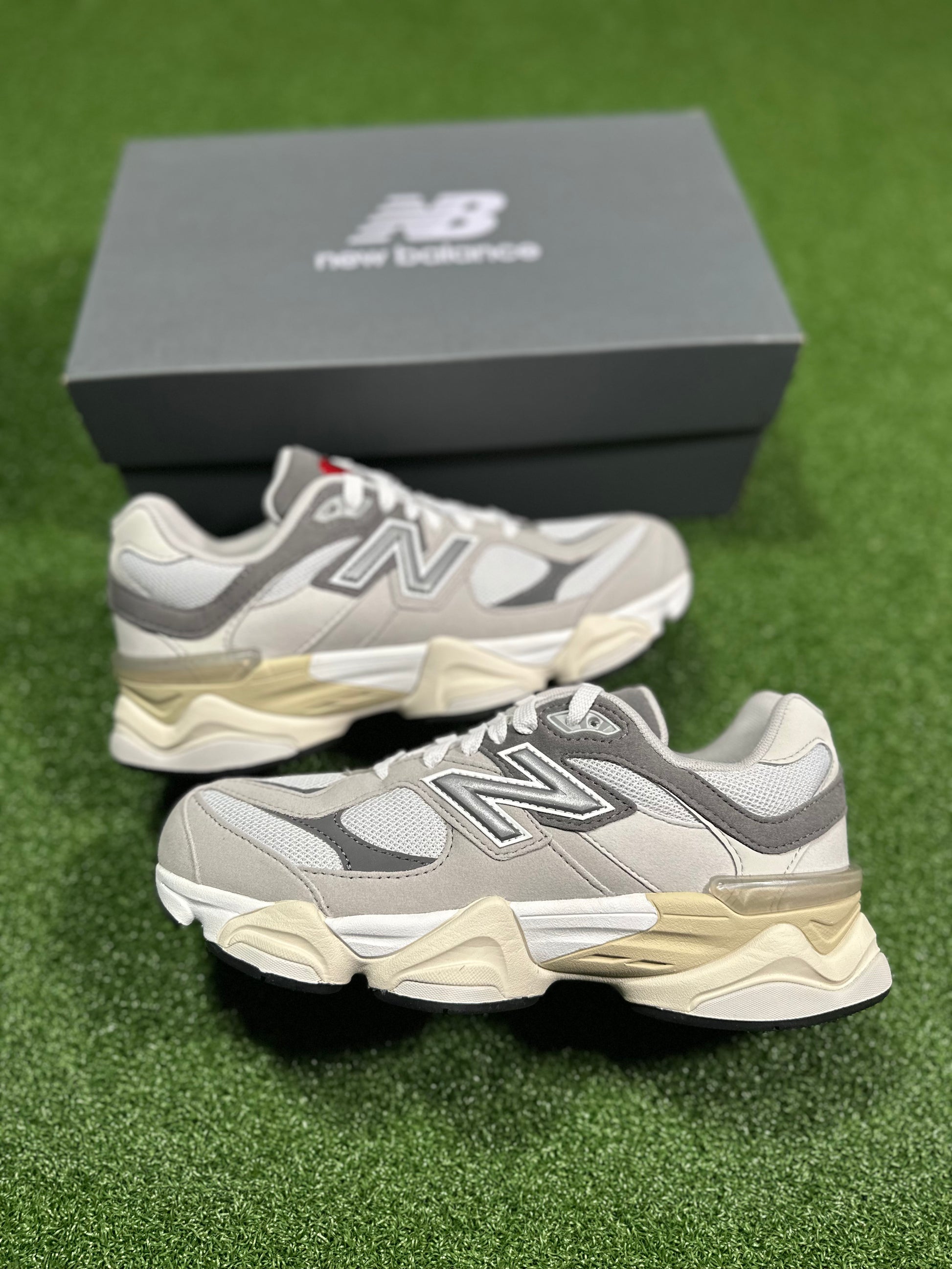 A pair of New Balance 9060 Rain Cloud sneakers for grade school students, displayed on a green surface with a New Balance branded box in the background.