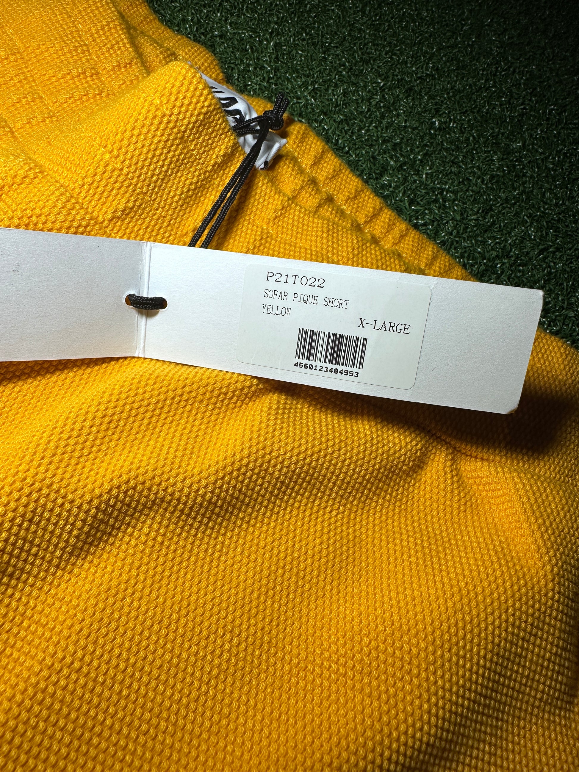 A close-up of a yellow textured garment with a white retail hangtag attached, photographed against a dark green textured background.