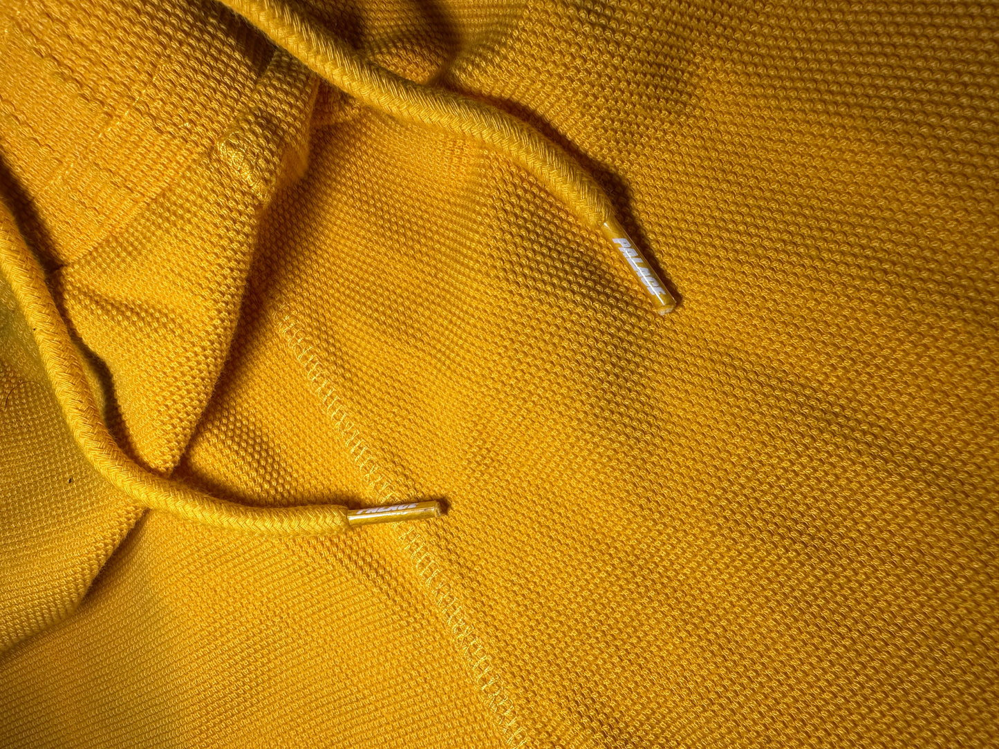 Close-up of a mustard yellow textured fabric, likely a knit hoodie or sweater, showing the drawstring cords with silver metal tips.