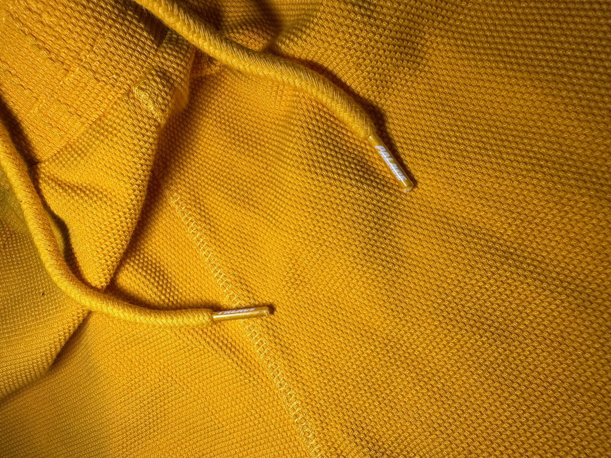 Close-up of a mustard yellow textured fabric, likely a knit hoodie or sweater, showing the drawstring cords with silver metal tips.