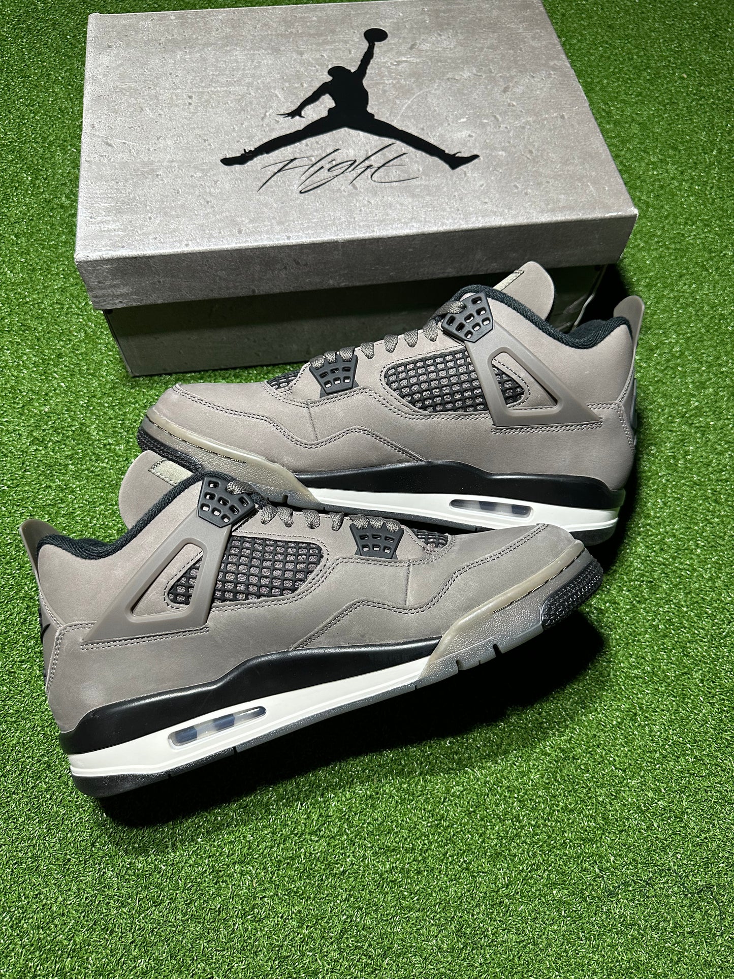 Gray sneakers with black accents on a green surface next to a Jordan box.Air Jordan 4 Retro Cave Stone suede leather sneaker side profile