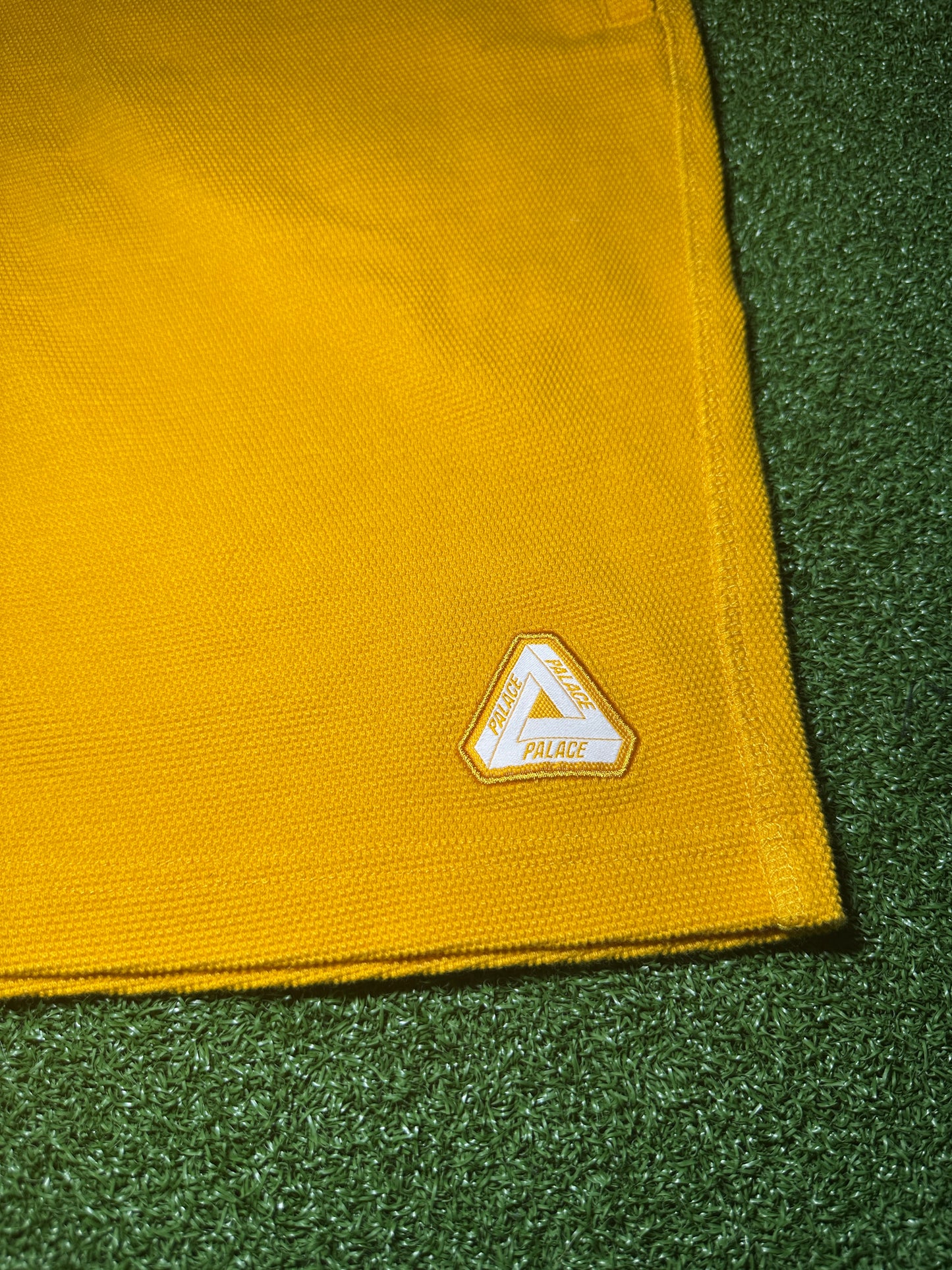 A close-up of a yellow woven fabric accessory, likely a golf towel or flag, featuring a white and gold triangular Palace logo patch, laid on a green turf-like surface.