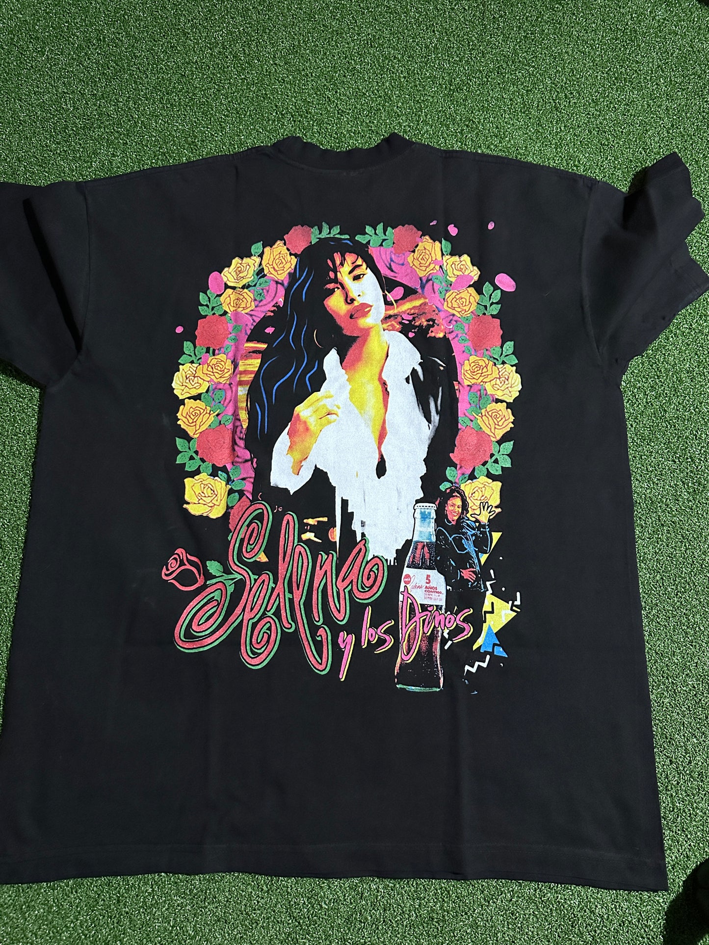 A black graphic t-shirt laid flat on green artificial turf. The shirt features a large, colorful graphic print of a woman surrounded by yellow and pink roses, with stylized pink script text on the right side.
