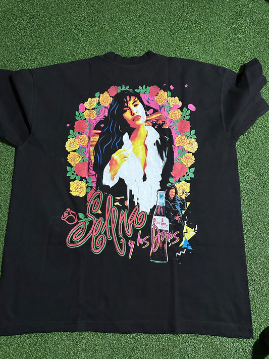 A black graphic t-shirt laid flat on green artificial turf. The shirt features a large, colorful graphic print of a woman surrounded by yellow and pink roses, with stylized pink script text on the right side.