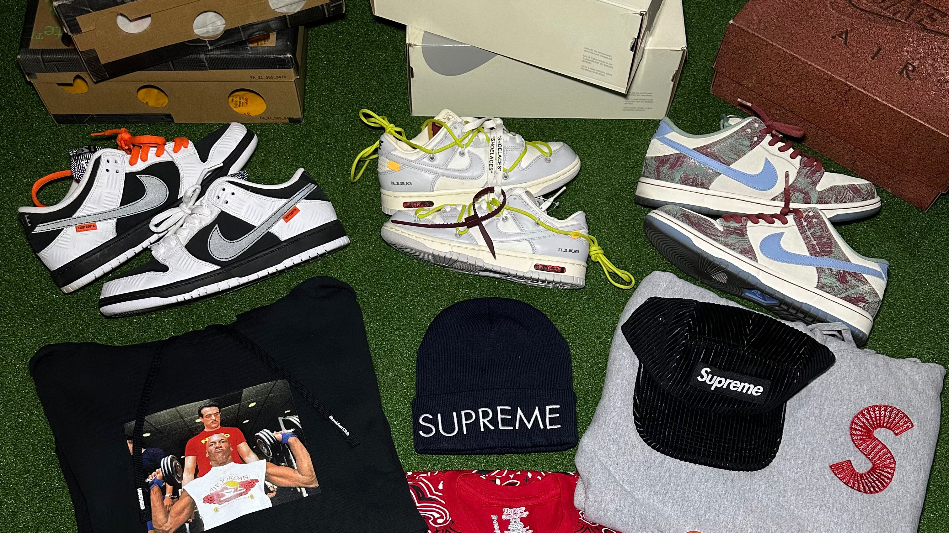 A flat lay collection of streetwear items on a green turf background, including Nike SB boxes and sneakers, Supreme apparel, and red and white sneakers.
